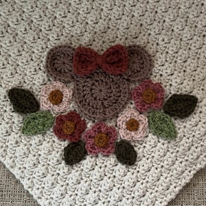 Può includere: Una copertina per neonati all'uncinetto di colore beige con un motivo floreale marrone e rosa. Il motivo presenta una testa di topo stilizzata con un fiocco rosso e un gruppo di fiori con foglie verdi.