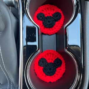 May include: Two red crochet car coasters with black Mickey Mouse silhouettes are placed in a car's cup holders. The coasters are round and made of a textured yarn. The car's interior is black and silver.