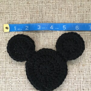 May include: A black crocheted Mickey Mouse head, approximately 4 inches wide, with two circular ears and a rounded head. The item is placed on a textured beige surface, with a blue measuring tape nearby.