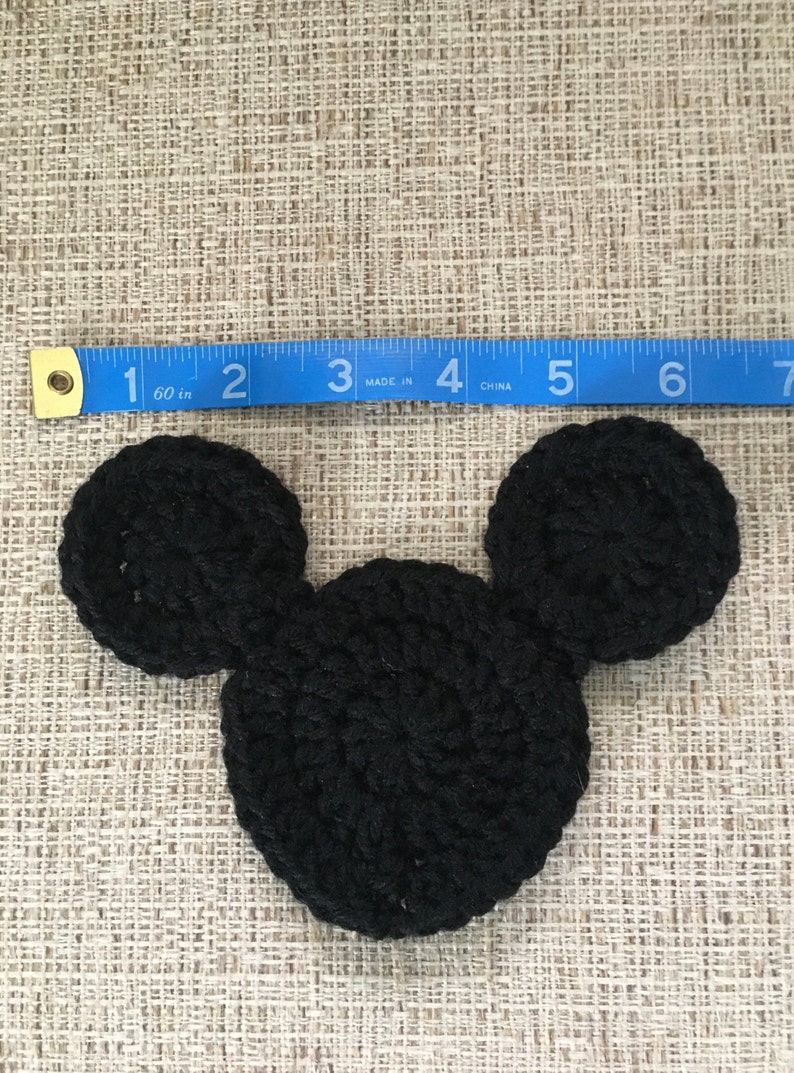 May include: A black crocheted Mickey Mouse head, approximately 4 inches wide, with two circular ears and a rounded head. The item is placed on a textured beige surface, with a blue measuring tape nearby.