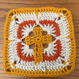 May include: A crocheted square with a cross design in the center. The square is made with yellow, white, and orange yarn.