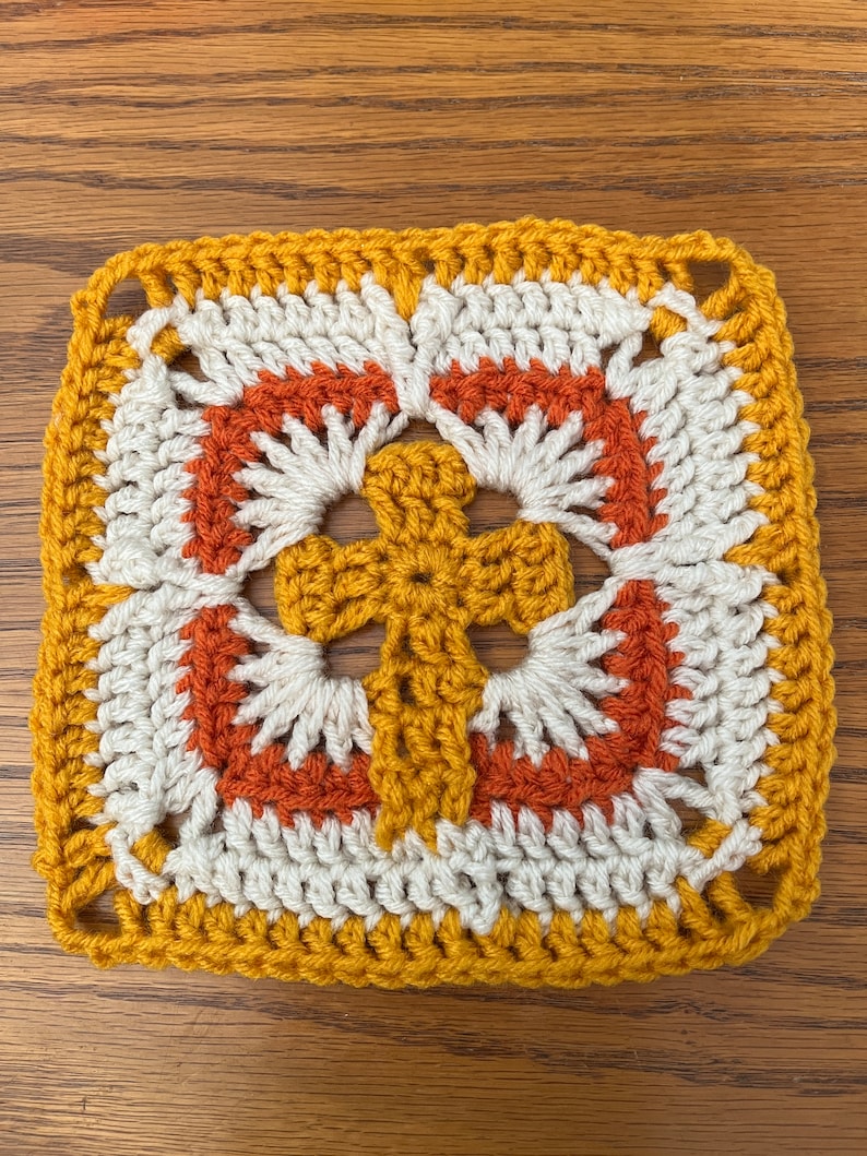 May include: A crocheted square with a cross design in the center. The square is made with yellow, white, and orange yarn.