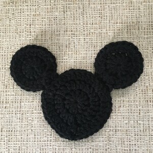 May include: A black, crocheted Mickey Mouse head shape, featuring a large circular head and two smaller circular ears. The handmade item is displayed on a textured, neutral-toned surface.