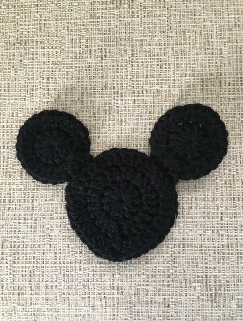 May include: A black, crocheted Mickey Mouse head shape, featuring a large circular head and two smaller circular ears. The handmade item is displayed on a textured, neutral-toned surface.