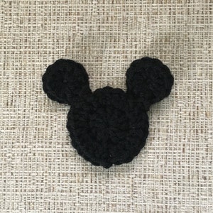 May include: A black crocheted Mickey Mouse head shape, featuring two rounded ears and a circular head. The handmade item is displayed against a textured, neutral-toned background, showcasing the detailed stitching of the yarn.
