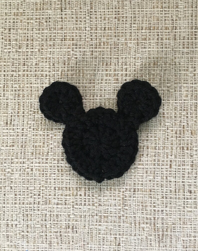 May include: A black crocheted Mickey Mouse head shape, featuring two rounded ears and a circular head. The handmade item is displayed against a textured, neutral-toned background, showcasing the detailed stitching of the yarn.
