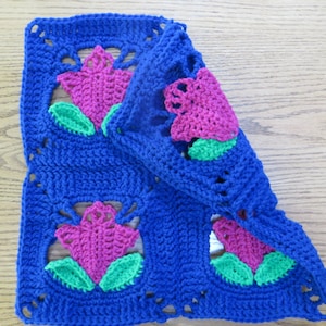 May include: A crocheted square with a blue background and pink and green flowers. The square is folded in half, showing the back side of the fabric.