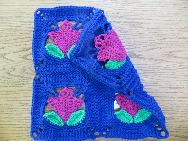May include: A crocheted square with a blue background and pink and green flowers. The square is folded in half, showing the back side of the fabric.
