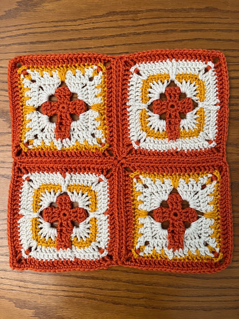 May include: Four crocheted squares in orange, white, and yellow, joined together to form a larger square. Each square features a central flower design.