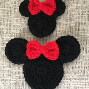 May include: Two black crochet mouse head shapes with red bows. The ears are round and the bows are centered on the head. The items are handmade and sit on a textured, neutral-colored surface.
