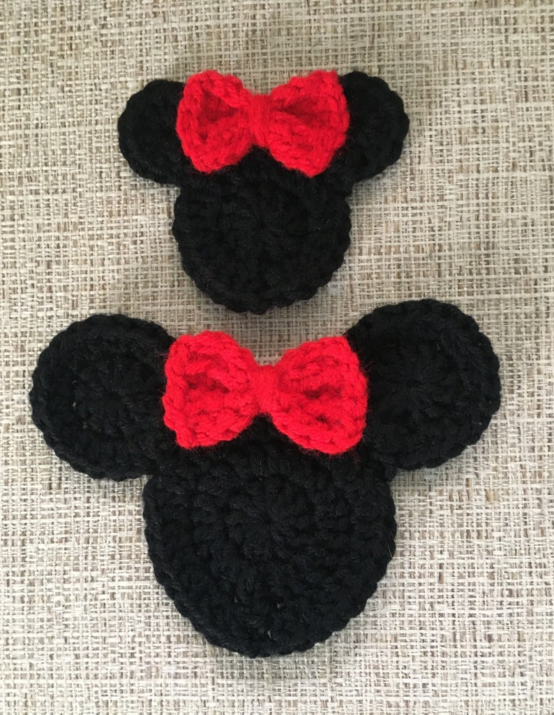 May include: Two black crochet mouse head shapes with red bows. The ears are round and the bows are centered on the head. The items are handmade and sit on a textured, neutral-colored surface.