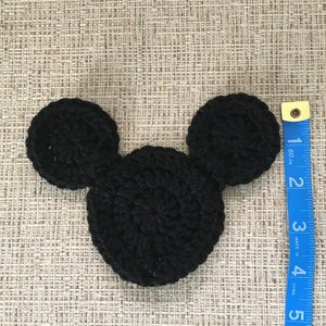 May include: A black crocheted Mickey Mouse head shape, approximately 4 inches wide, lies on a textured beige surface. A blue measuring tape is positioned to the right, showing inches. The handmade item is likely a craft supply or decorative element.