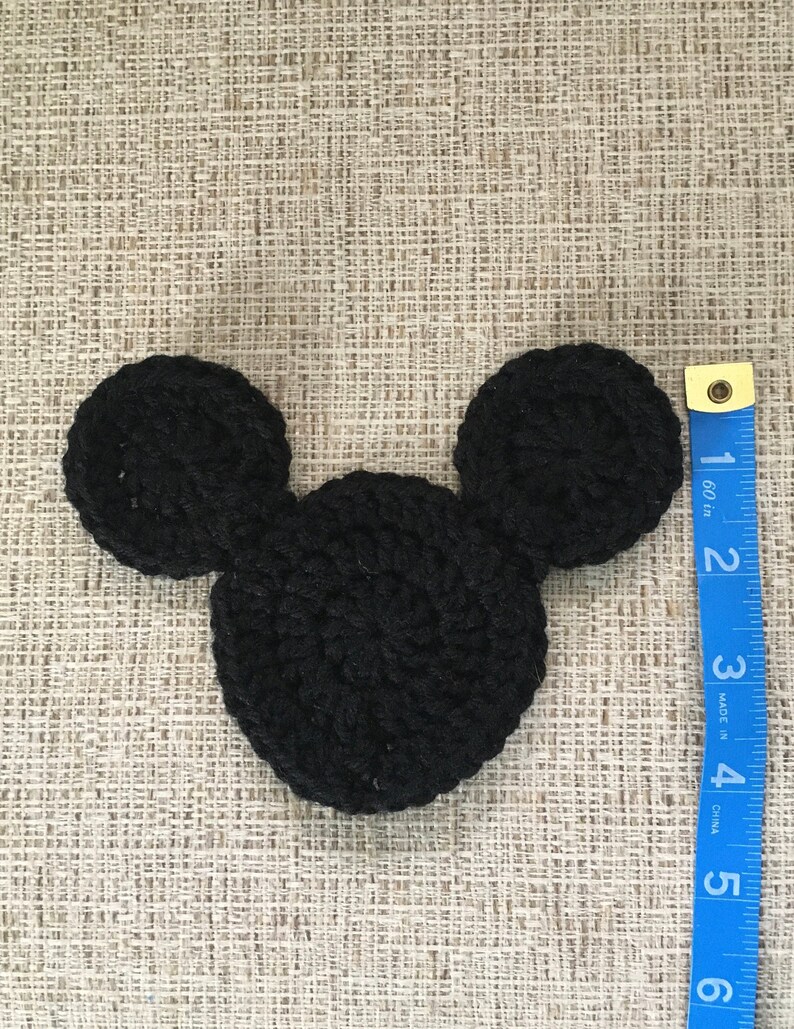 May include: A black crocheted Mickey Mouse head shape, approximately 4 inches wide, lies on a textured beige surface. A blue measuring tape is positioned to the right, showing inches. The handmade item is likely a craft supply or decorative element.