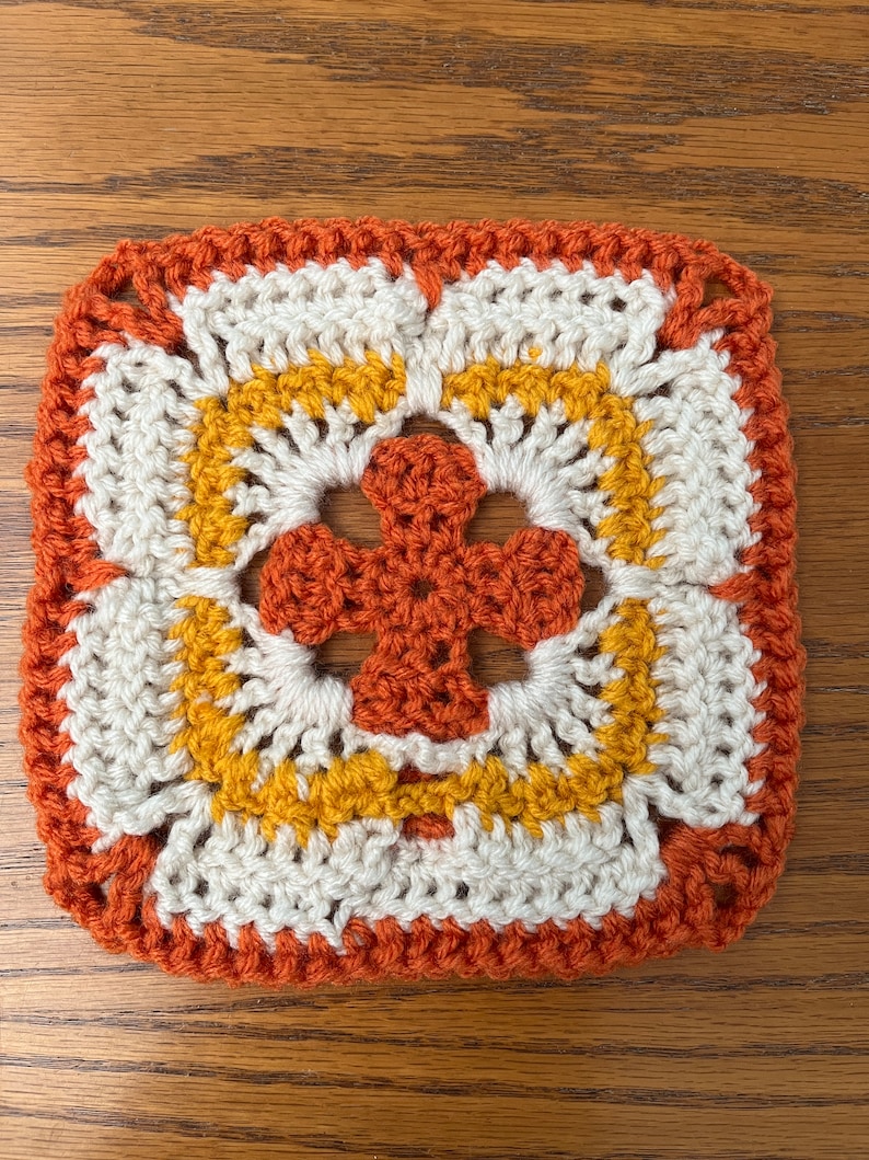 May include: A crocheted square with a flower design in orange, yellow, and white.