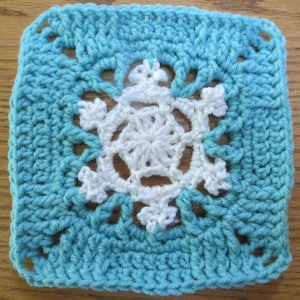 May include: A crocheted square with a white snowflake design in the center. The square is a light blue color.