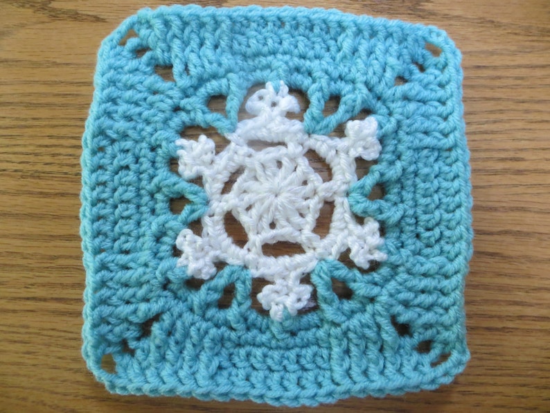 May include: A crocheted square with a white snowflake design in the center. The square is a light blue color.
