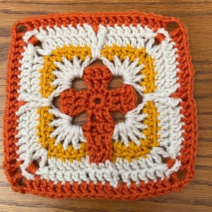 May include: A crocheted square with a cross design in the center. The cross is made with orange yarn, and the square is made with white, orange, and yellow yarn.