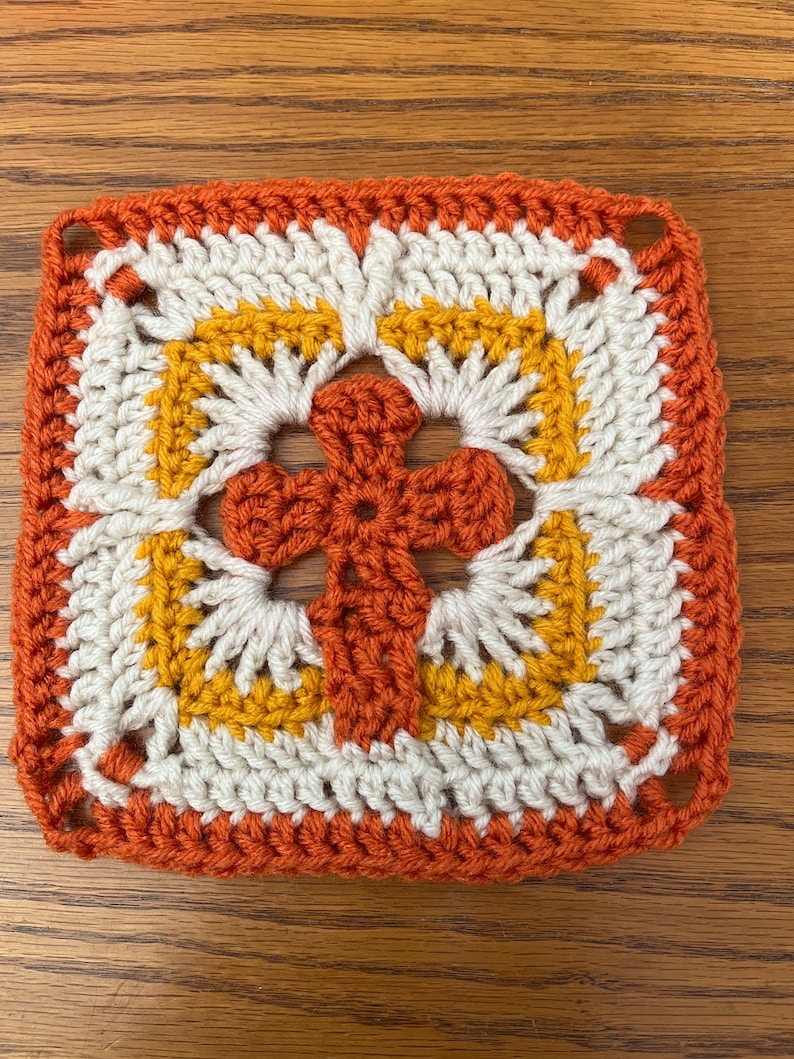 May include: A crocheted square with a cross design in the center. The cross is made with orange yarn, and the square is made with white, orange, and yellow yarn.