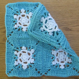 May include: A crocheted square with a blue background and white snowflake designs. The square is folded in half, showing the back side of the fabric.