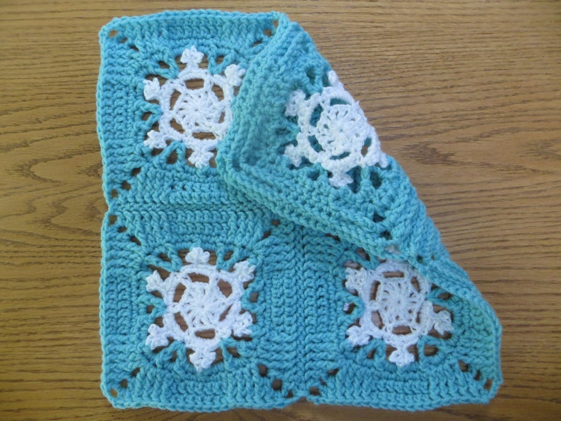 May include: A crocheted square with a blue background and white snowflake designs. The square is folded in half, showing the back side of the fabric.