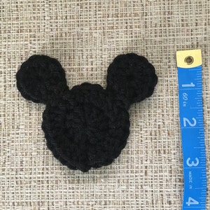 May include: A black crocheted Mickey Mouse head shape, approximately 3 inches wide, lies on a textured beige surface. The ears are rounded and attached to a circular head. A blue measuring tape is visible.