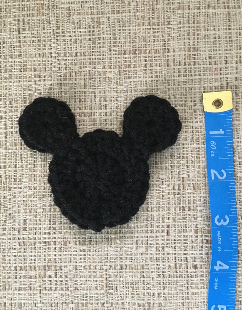 May include: A black crocheted Mickey Mouse head shape, approximately 3 inches wide, lies on a textured beige surface. The ears are rounded and attached to a circular head. A blue measuring tape is visible.