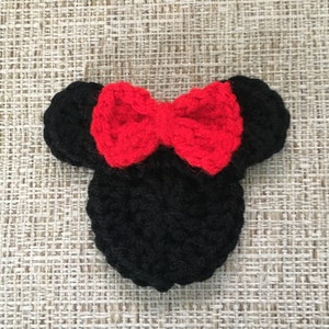 May include: A handmade black crochet mouse head with a bright red bow. The ears are round and the bow is centered on the head. The item is likely a decorative piece or accessory.
