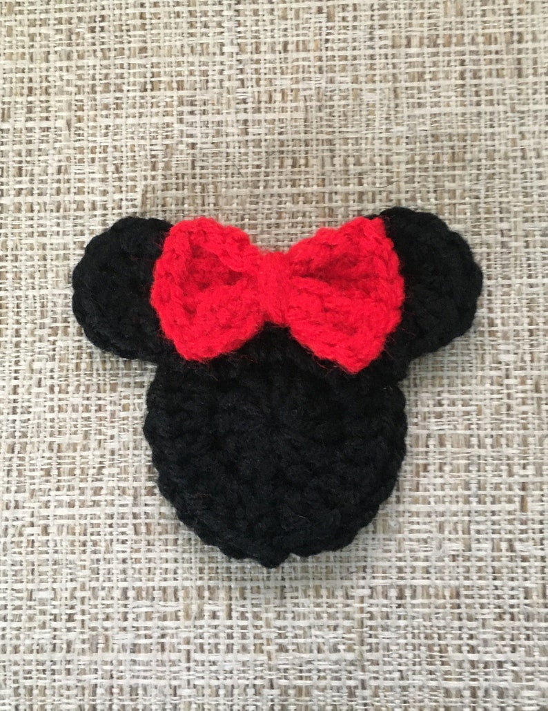 May include: A handmade black crochet mouse head with a bright red bow. The ears are round and the bow is centered on the head. The item is likely a decorative piece or accessory.