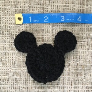 May include: A black crocheted Mickey Mouse head, approximately 2 inches wide, lies on a textured beige surface. The ears are rounded and attached to a circular head. A blue measuring tape is placed above the crocheted item.