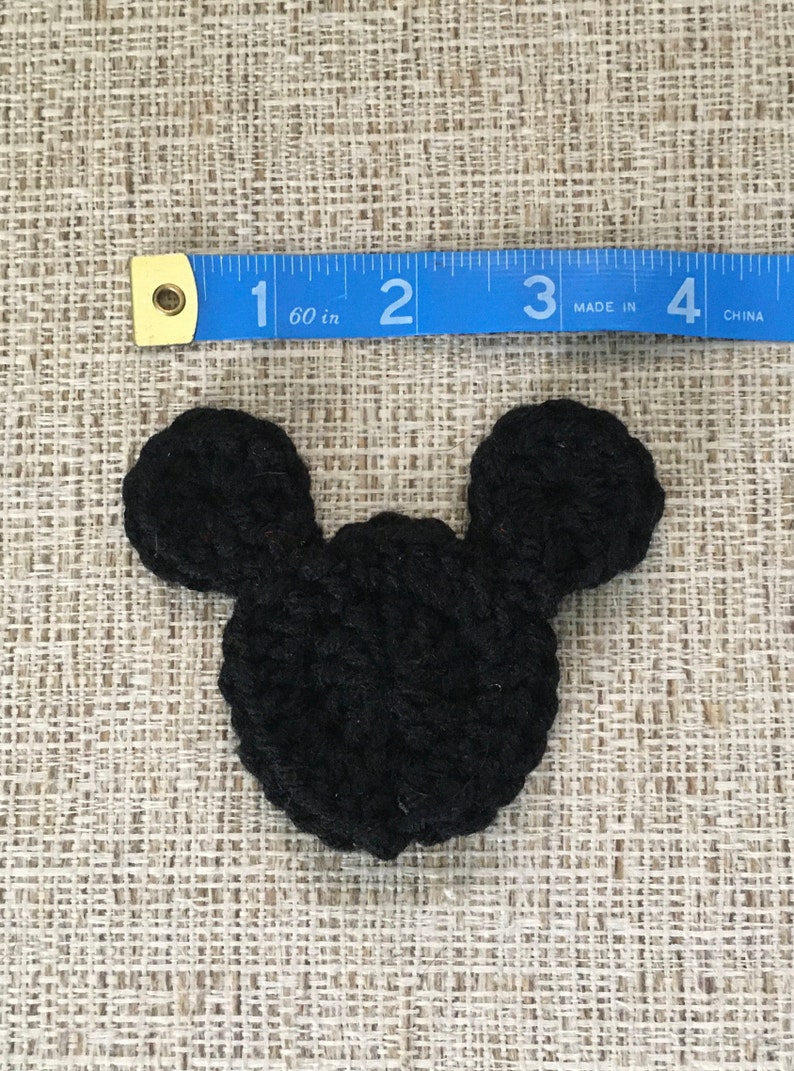 May include: A black crocheted Mickey Mouse head, approximately 2 inches wide, lies on a textured beige surface. The ears are rounded and attached to a circular head. A blue measuring tape is placed above the crocheted item.