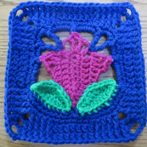 May include: A crocheted square with a pink flower and green leaves in the center. The square is surrounded by a blue border.