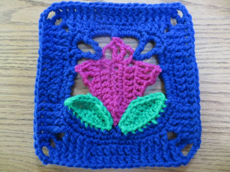 May include: A crocheted square with a pink flower and green leaves in the center. The square is surrounded by a blue border.