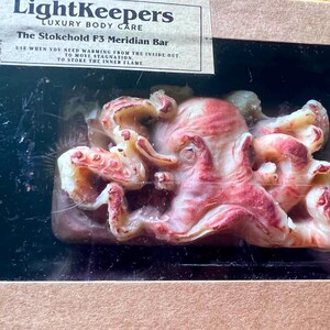 May include: A bar of soap in a box, featuring a detailed octopus design with red and cream tones. The soap is labeled "The Stokehold F3 Meridian Bar" from LightKeepers Luxury Body Care. The packaging is a dark brown box.