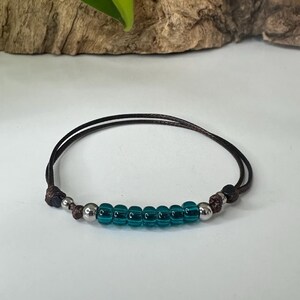 May include: A handmade bracelet featuring a dark brown cord with teal and silver beads. The bracelet has a simple design with a row of teal beads in the center, flanked by silver and brown beads. The background is a white surface with a wooden accent.