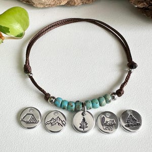 May include: A brown cord bracelet with turquoise beads and silver accents. Five silver charms with camping, mountain, tree, raccoon, and campfire designs are displayed below the bracelet. The bracelet is on a white surface.