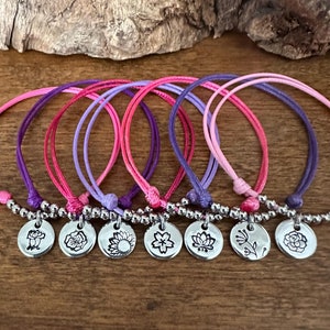 May include: Set of six colorful friendship bracelets with silver charms featuring different flower designs. The bracelets are made with pink, purple, and lavender cord.