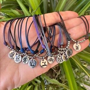 K&ouml;nnte beinhalten: Eine Sammlung von verstellbaren Kordelarmb&auml;ndern mit silberfarbenen runden Anh&auml;ngern. Jeder Anh&auml;nger zeigt ein einzigartiges eingraviertes Motiv, darunter ein Hirsch, ein UFO, eine Kreaturensilhouette und eine Fee. Die Armb&auml;nder sind mit kleinen Silberperlen besetzt.