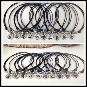 K&ouml;nnte beinhalten: Eine Sammlung von zehn verstellbaren Kordelarmb&auml;ndern in verschiedenen dunklen Farben, jedes mit einem runden silberfarbenen Metallanh&auml;nger. Die Anh&auml;nger sind mit markanten schwarzen Silhouetten von Fabelwesen, Tieren und einer fliegenden Untertasse graviert.
