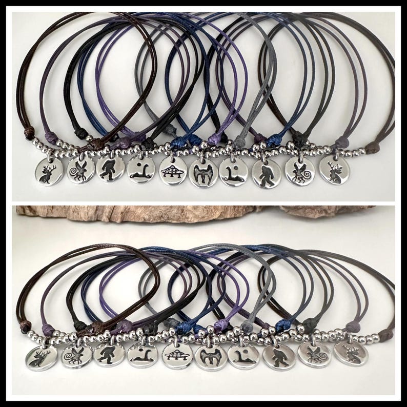 K&ouml;nnte beinhalten: Eine Sammlung von zehn verstellbaren Kordelarmb&auml;ndern in verschiedenen dunklen Farben, jedes mit einem runden silberfarbenen Metallanh&auml;nger. Die Anh&auml;nger sind mit markanten schwarzen Silhouetten von Fabelwesen, Tieren und einer fliegenden Untertasse graviert.