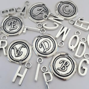 May include: A collection of silver-toned metal letter charms with a variety of styles, including plain letters, letters with a wax seal design, and letters with a decorative border. The charms have jump rings for attaching to jewelry or other crafts.