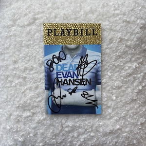 Könnte beinhalten: Ein "Dear Evan Hansen"-Programmheft mit goldenem Glitzer oben und Unterschriften. Das Programmheft zeigt ein blau-weiß gestreiftes Hemd und den Text "DEAR EVAN HANSEN". Die Unterschriften sind in schwarzer Tinte.