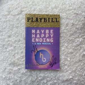 May include: A Playbill for the musical "Maybe Happy Ending" with a gold glitter top. The cover is purple with the title in white and the words "A New Musical". The Playbill has signatures on the front.