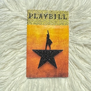 May include: A decorative playbill with the word "PLAYBILL" in gold glitter at the top. The cover features a silhouette of a person on a black star, set against an orange and yellow gradient background.