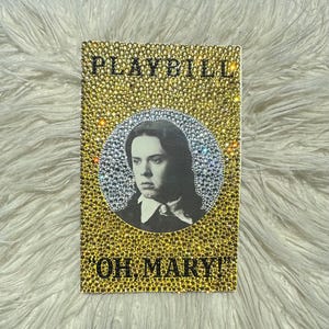 May include: A rectangular playbill with a portrait of a person in a black and white circle. The playbill is covered in gold and silver rhinestones. The words "PLAYBILL" and "OH, MARY!" are written in gold rhinestones.