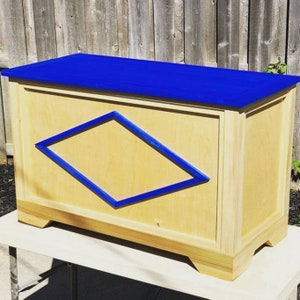 May include: A wooden toy chest with a blue lid and a blue diamond shape on the front. The chest is sitting on a white table.