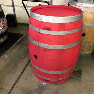 May include: A red wooden barrel with silver metal bands. The barrel is sitting on a concrete floor.