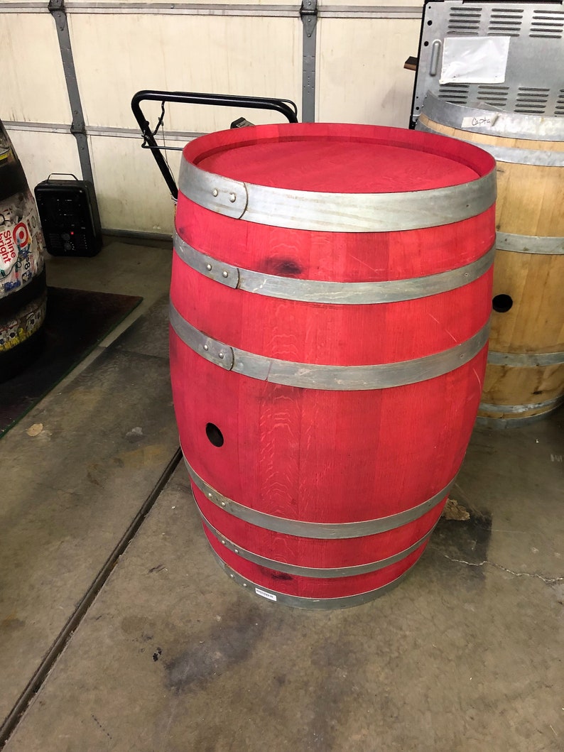 May include: A red wooden barrel with silver metal bands. The barrel is sitting on a concrete floor.