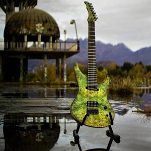 May include: A green and yellow electric guitar with a black fretboard and black hardware. The guitar is on a stand and is in front of a blurry background of a building and trees.