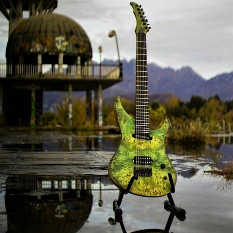 May include: A green and yellow electric guitar with a black fretboard and black hardware. The guitar is on a stand and is in front of a blurry background of a building and trees.