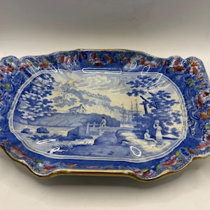 May include: A rectangular, antique blue and white ceramic serving tray with a landscape scene. The tray has a scalloped edge with floral accents and a gold trim. The central image depicts a coastal scene with figures and ships.